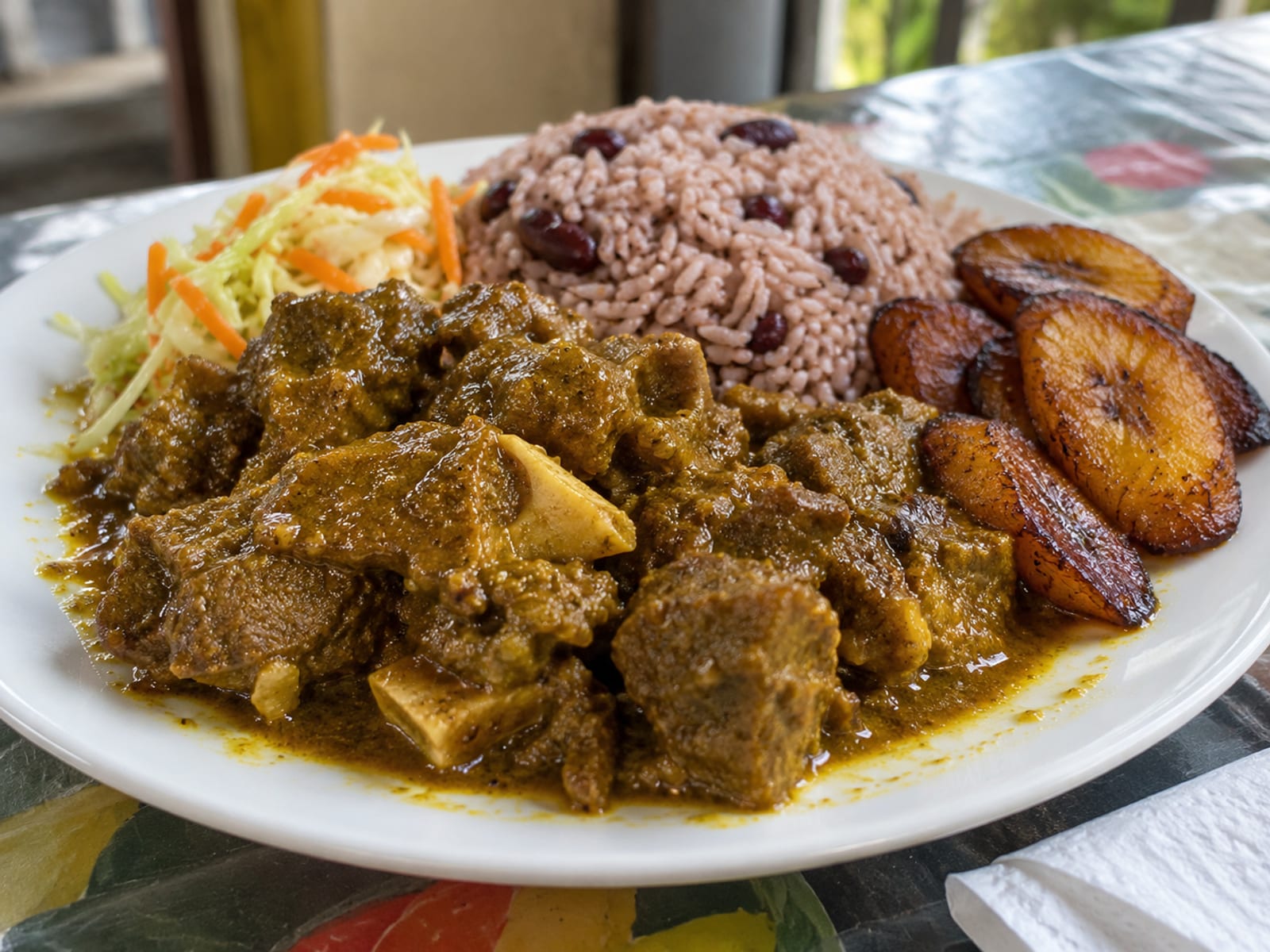 A plate of curry goat with rice and peas, fried plantain and a small salad