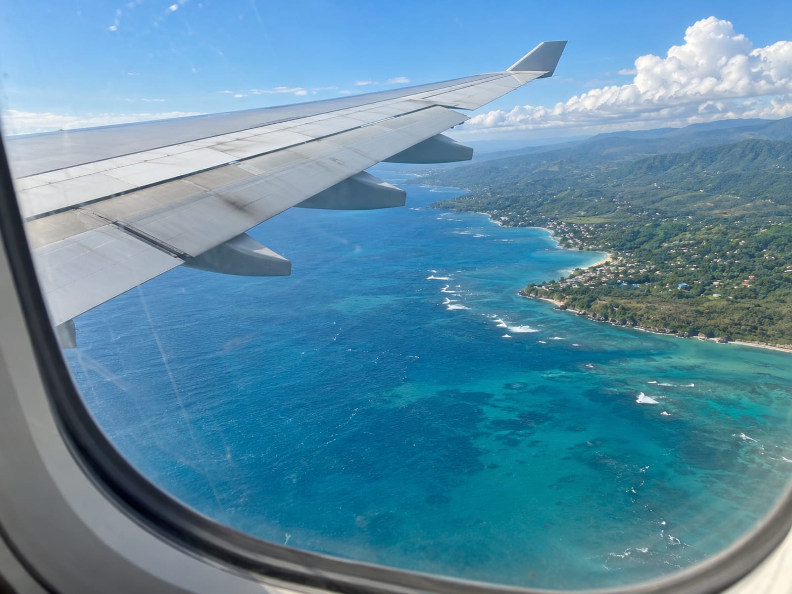 Passenger jet wing above turquoise Caribbean water and a green coastline