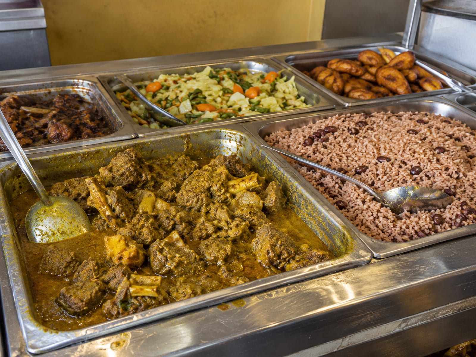 Steam trays of curry goat, rice and peas and plantain on a kitchen counter
