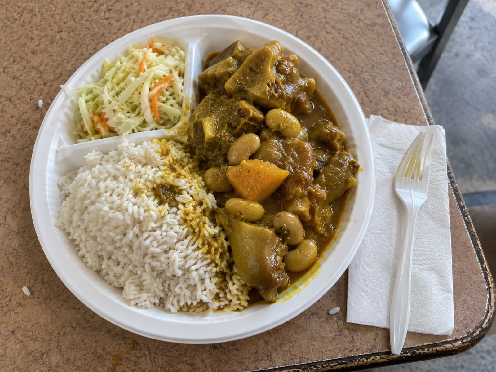 A plate of rice with a ladle of curried cow foot and beans on a plain lunch table