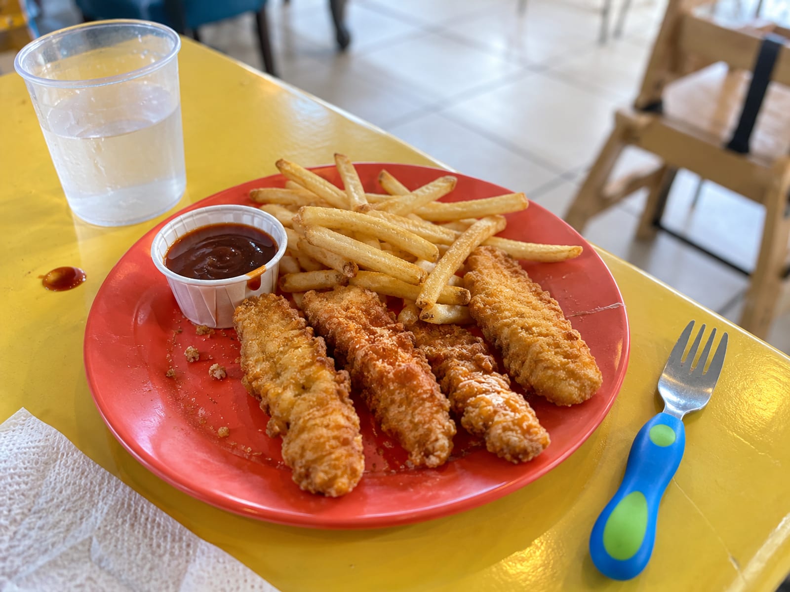 A children's plate of chicken fingers with fries and a small cup of sauce on a bright table
