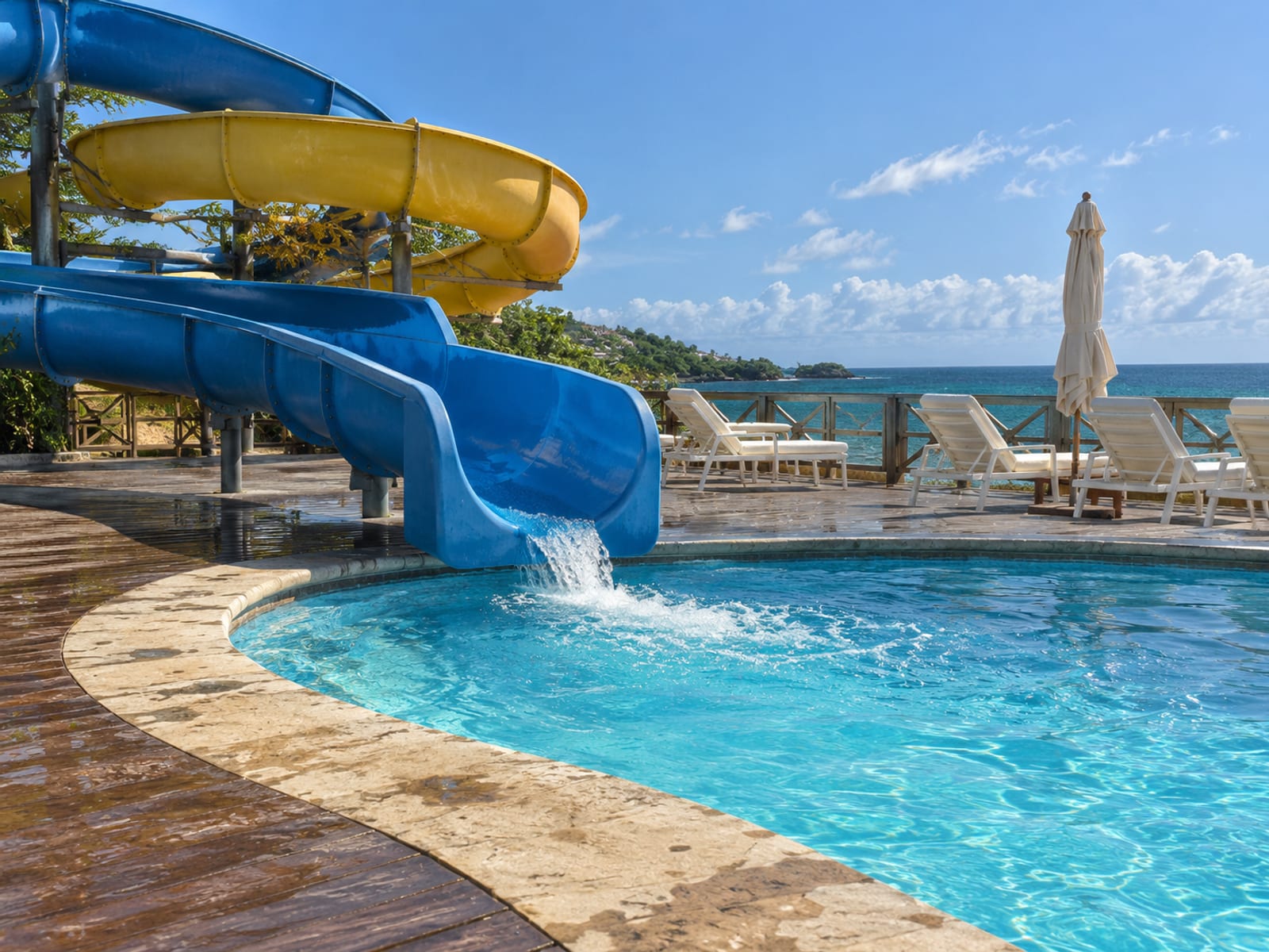A sunlit waterslide and pool deck beside the sea with empty lounge chairs