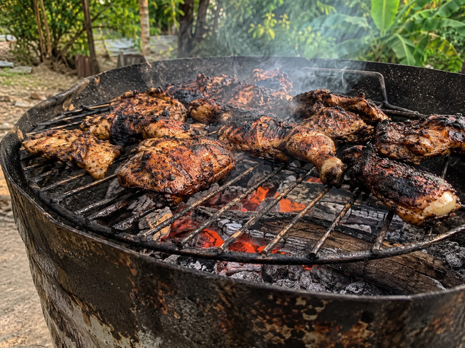 A steel jerk drum with smoke rising over glowing charcoal and pimento wood