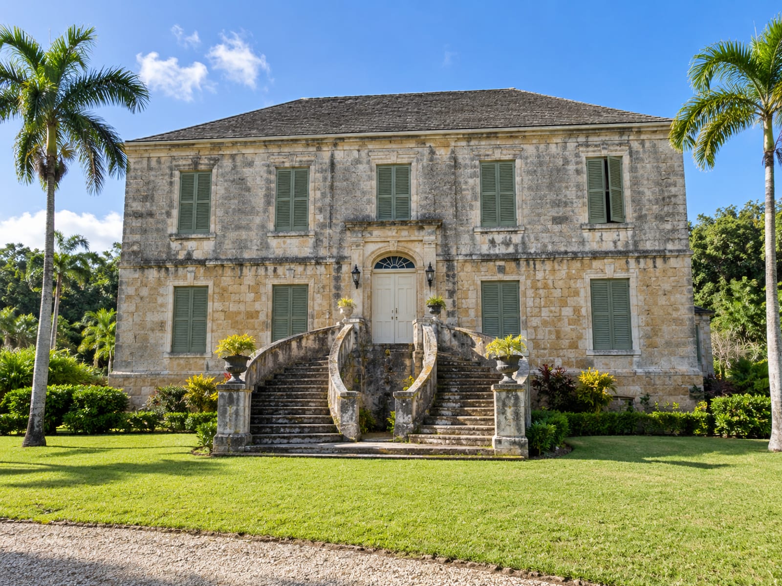 A stately two-storey stone mansion with a broad staircase and palm trees under blue sky