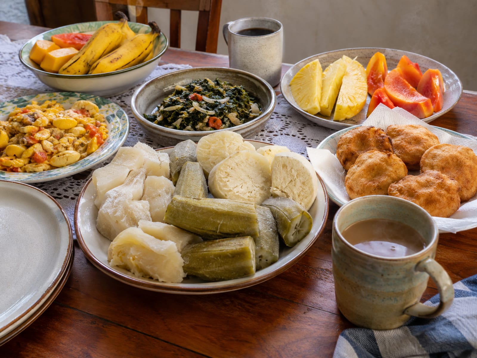 Breakfast table set with boiled bananas, yam, fried dumplings, fruit and hot drinks
