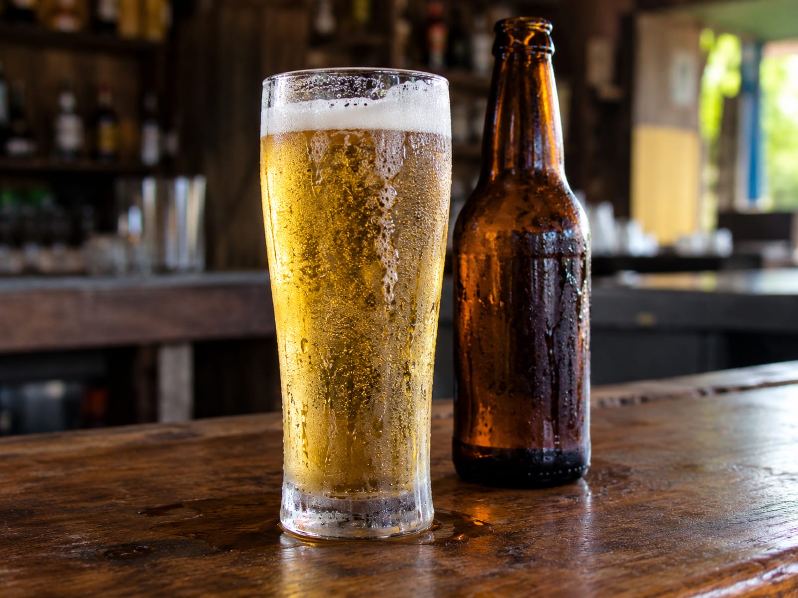 A tall glass of golden lager beside an unlabelled bottle on a bar counter