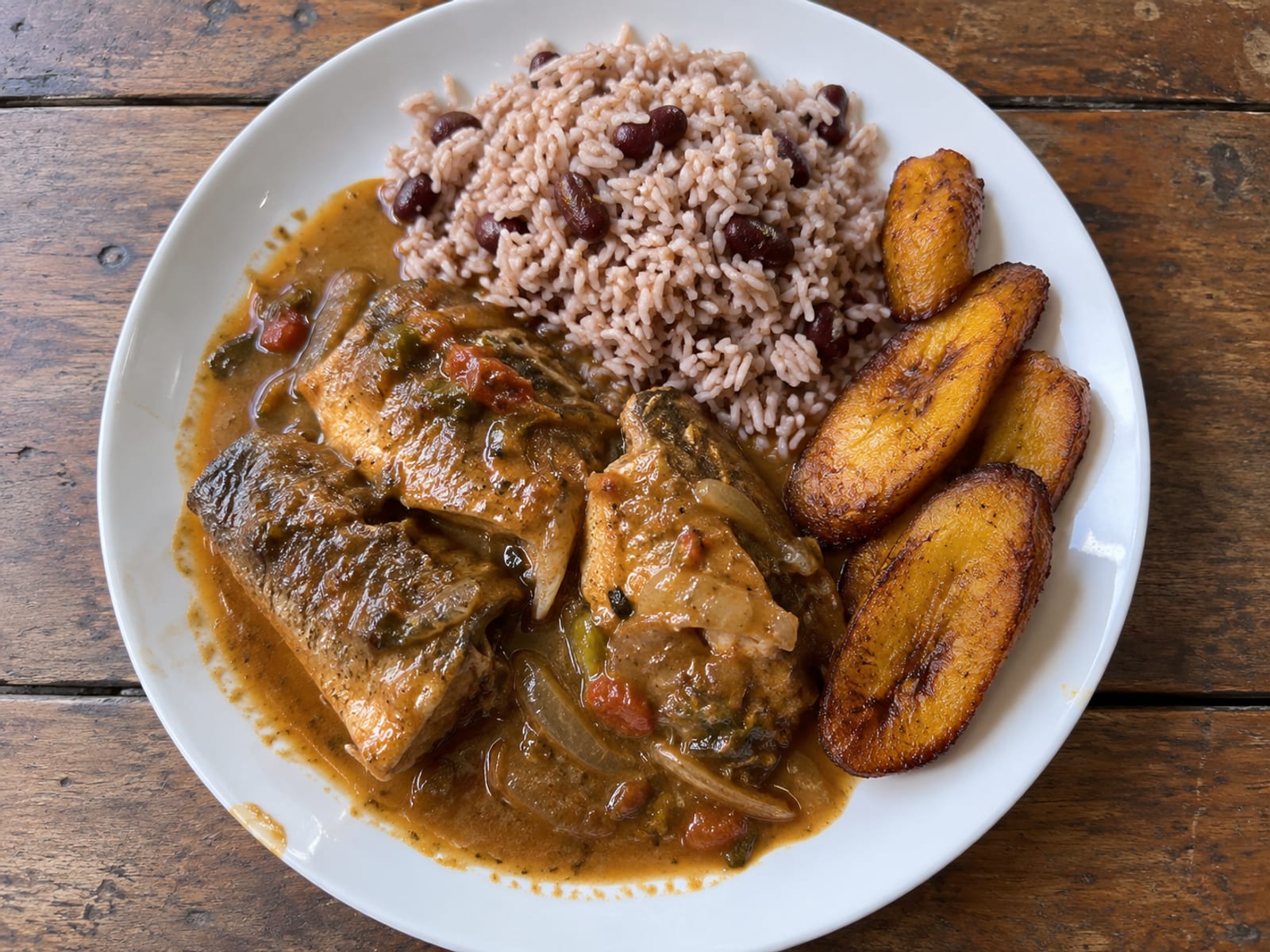 Plate of brown stewed fish with rice and peas and fried plantain on a wooden table