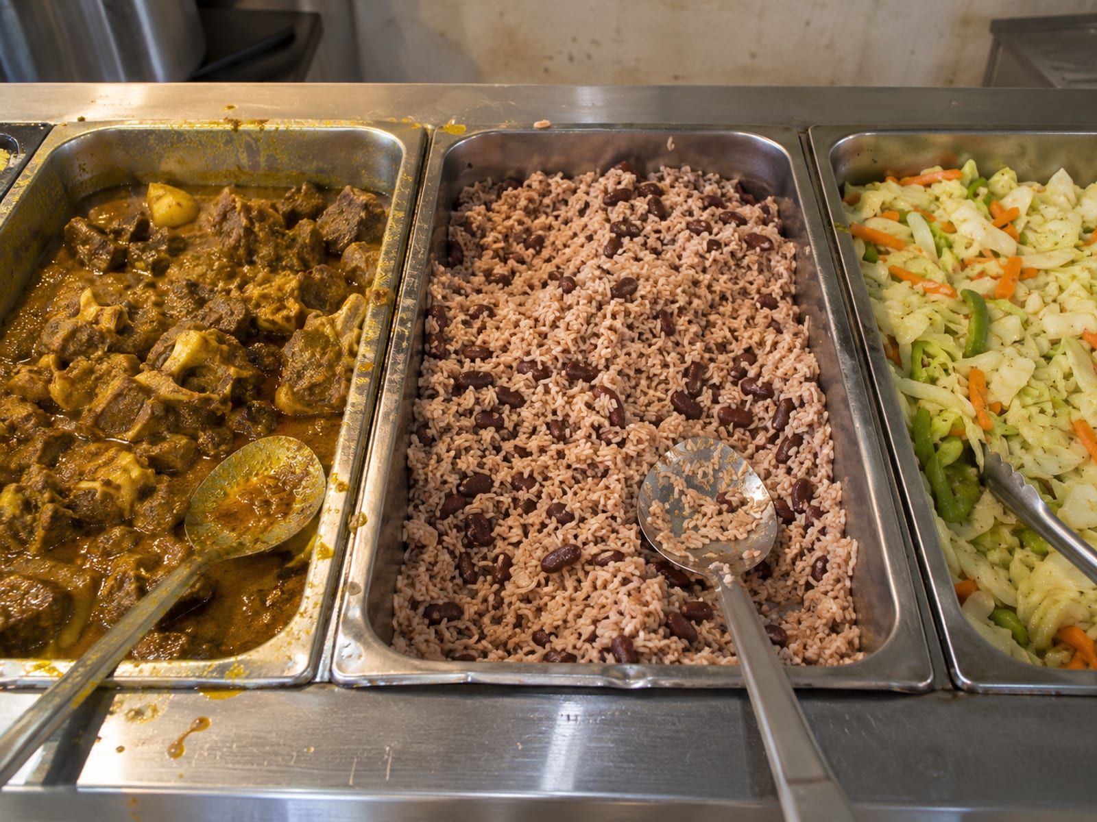 Steam trays of curry goat, rice and peas and cabbage behind a plain counter