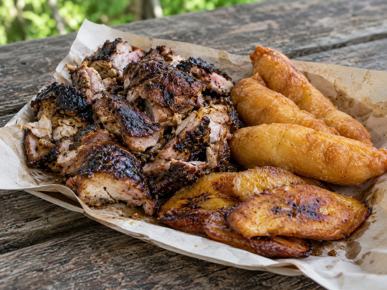 Jerk chicken, festival and fried plantain on a paper-lined tray on a picnic table