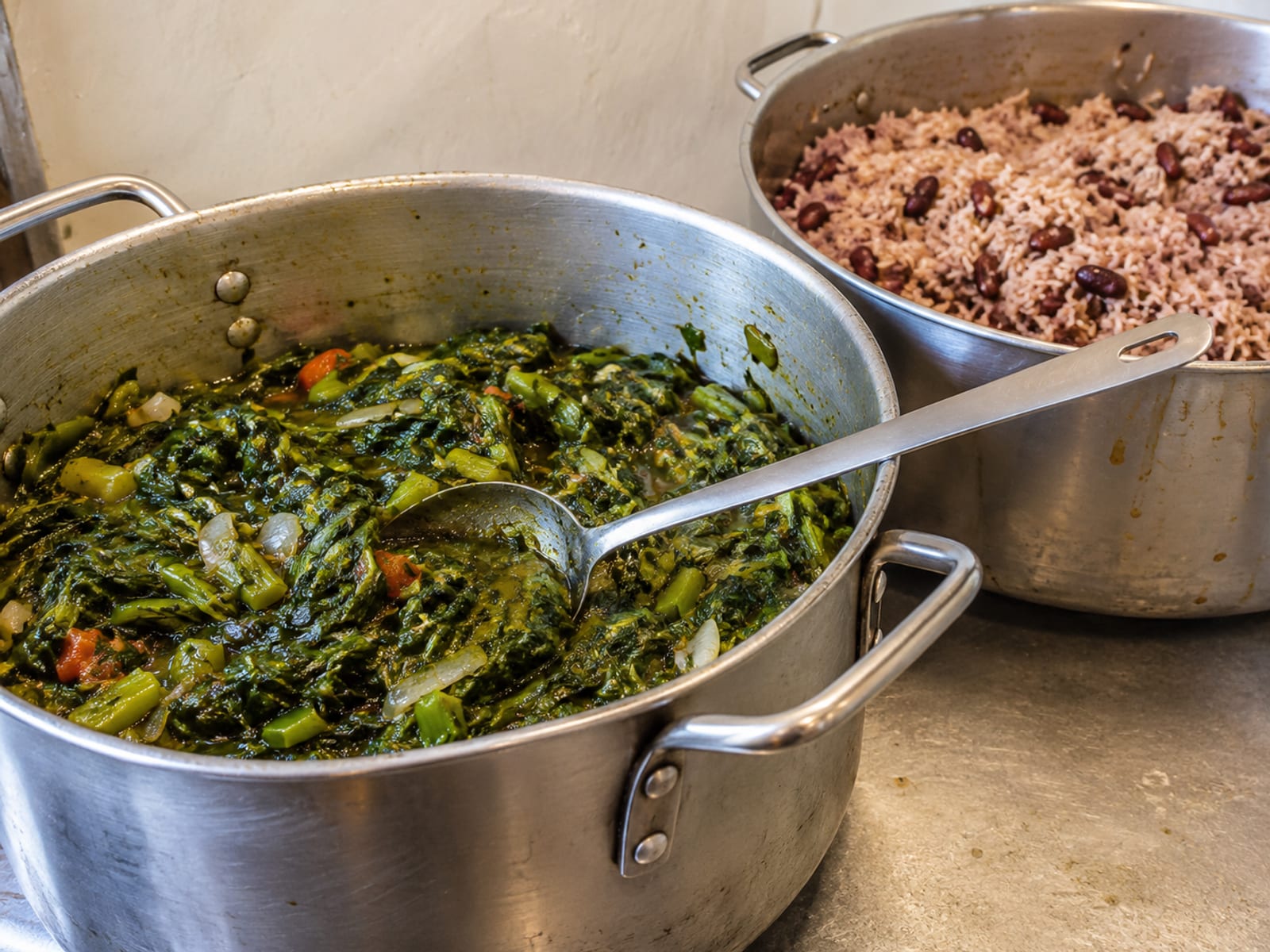 Pots of callaloo and rice and peas with a ladle on a cookshop counter
