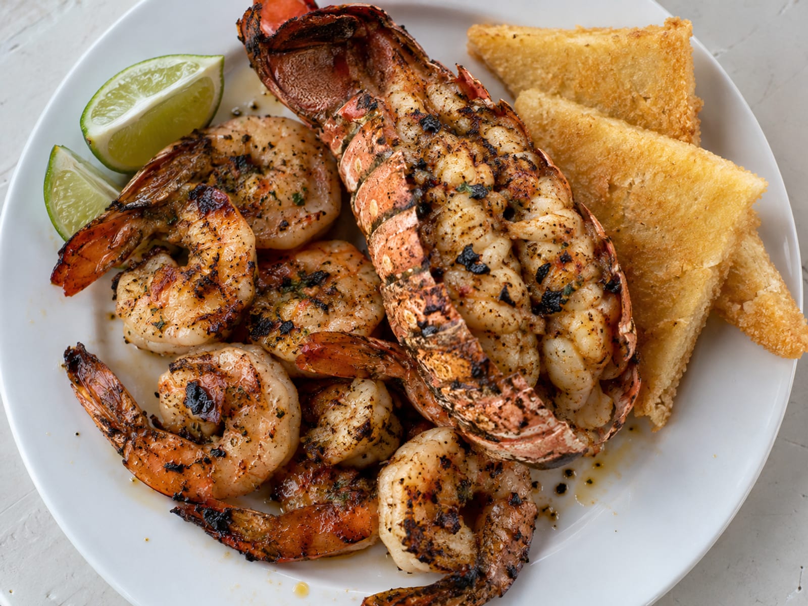 Grilled shrimp and a lobster tail on a white plate with lime wedges and bammy